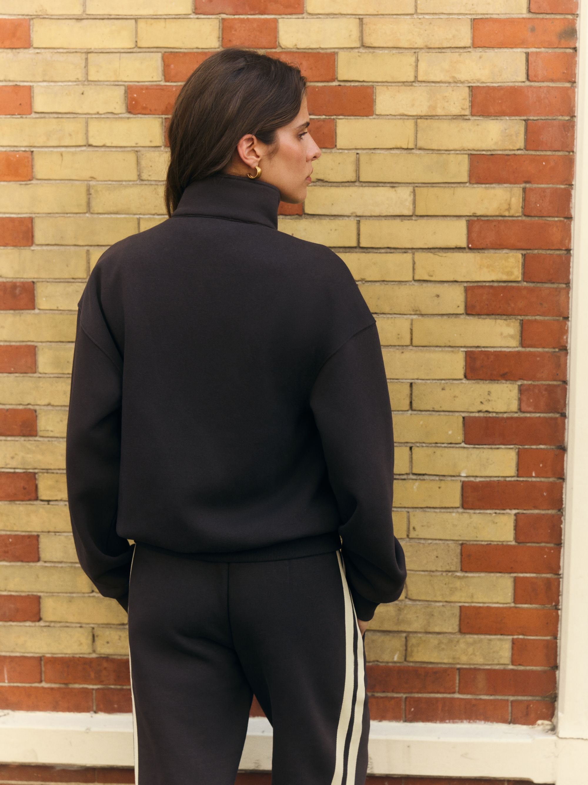 Woman from behind wearing an espresso CASPARA funnel zip sweatshirt against a brick wall.